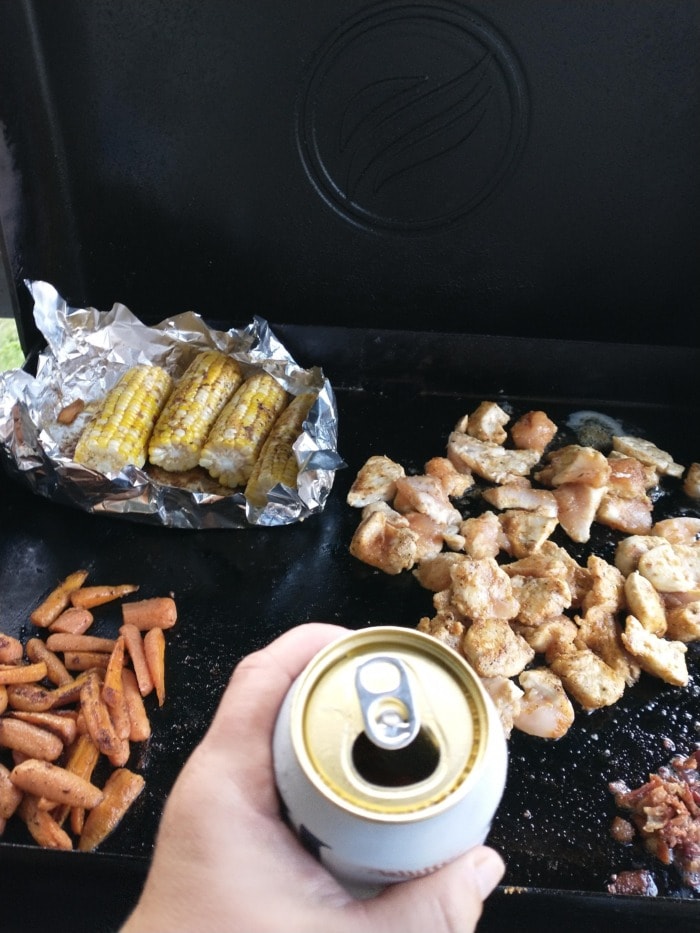 a hand holding an alcohol free beer for cooking in front of a barbecue with vegetables and meat cuts