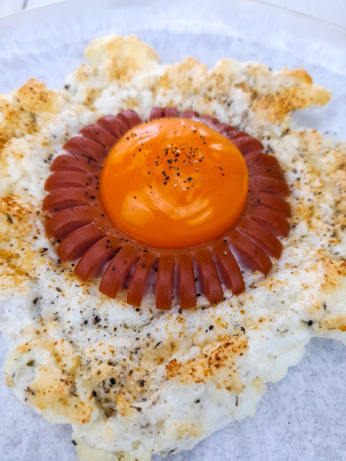A close up of a baked sunny side up egg with puffy egg white and a vienna sausage around the egg yolk 