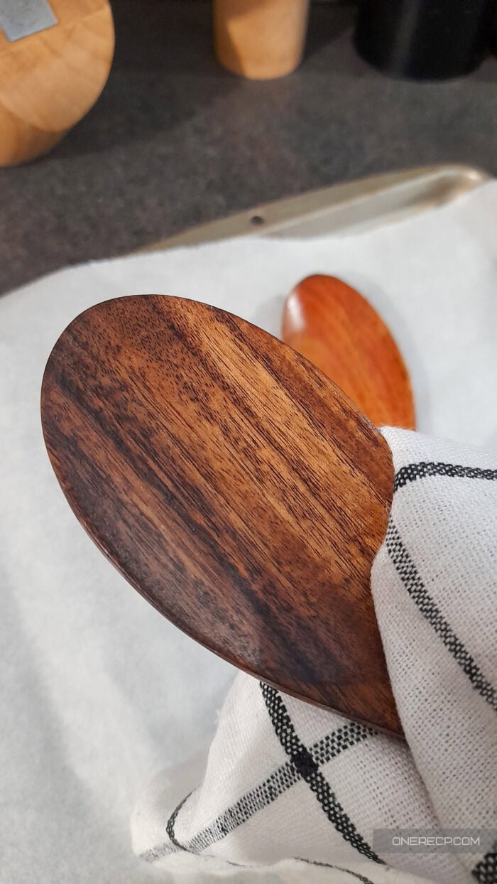 A person gently buffs a dark wooden spoon with a soft kitchen cloth to remove excess oil after seasoning.