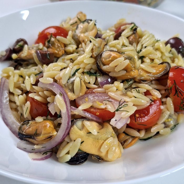 Mussel and orzo salad on a white plate with cherry tomatoes, Kalamata olives, red onion, and dill