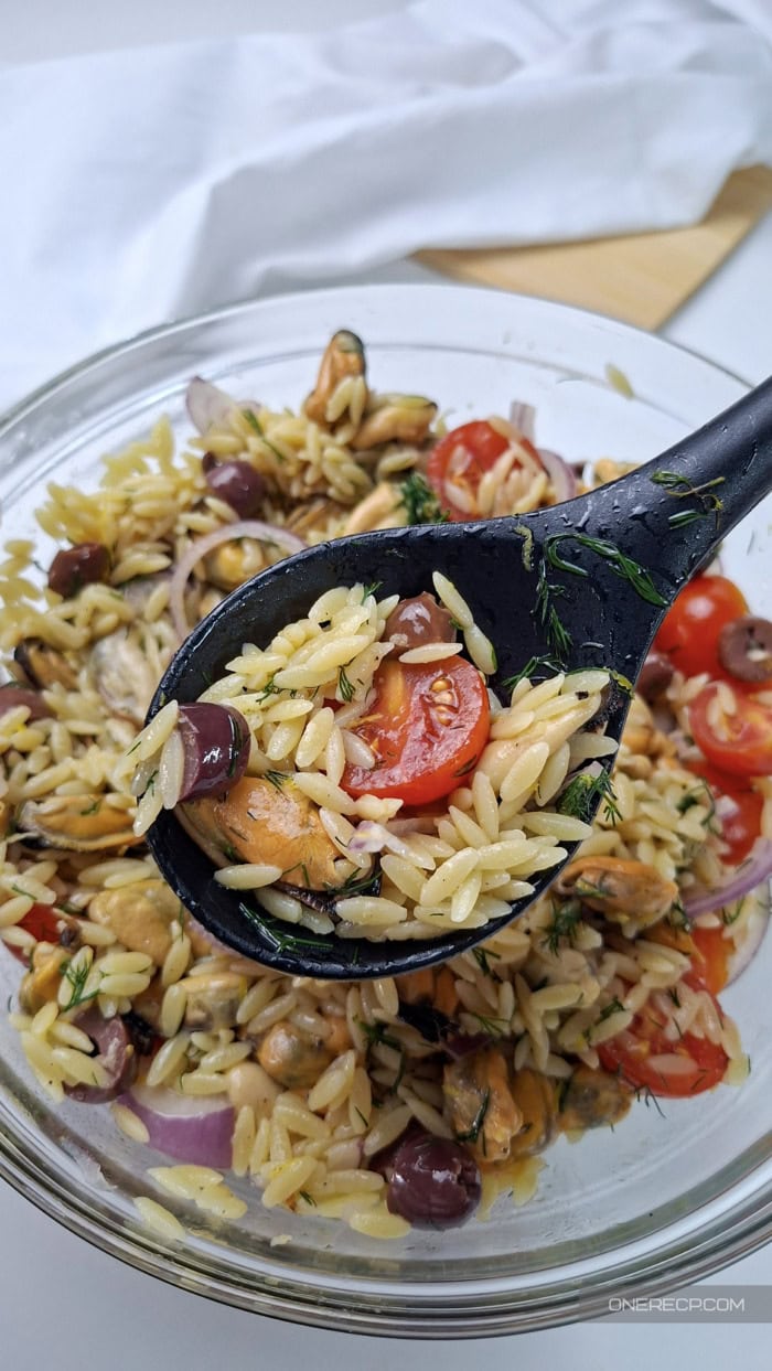 Spoonful of mussel and orzo salad with cherry tomato, Kalamata olive, red onion, and dill above a glass bowl