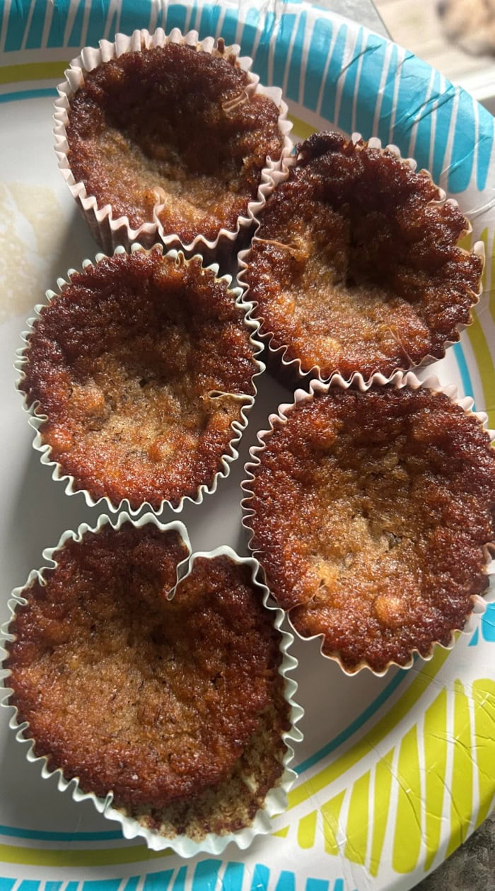 Five collapsed banana muffins in paper liners, showing sunken tops and dark caramelized edges on a colorful paper plate.