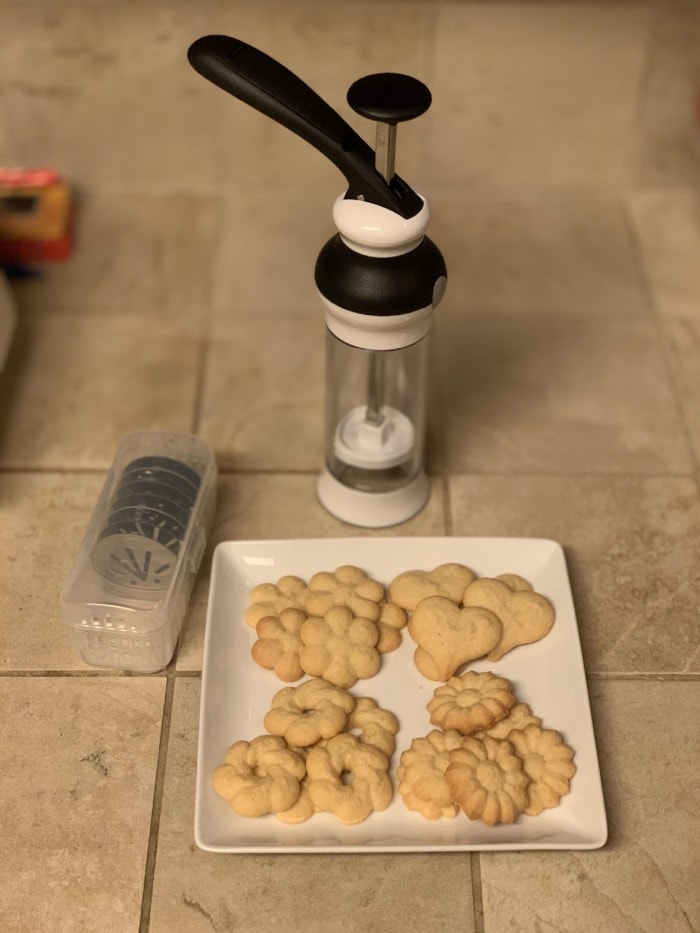 а cookie press behind a plate of spritz cookies, and a plastic box with press discs besides the plate