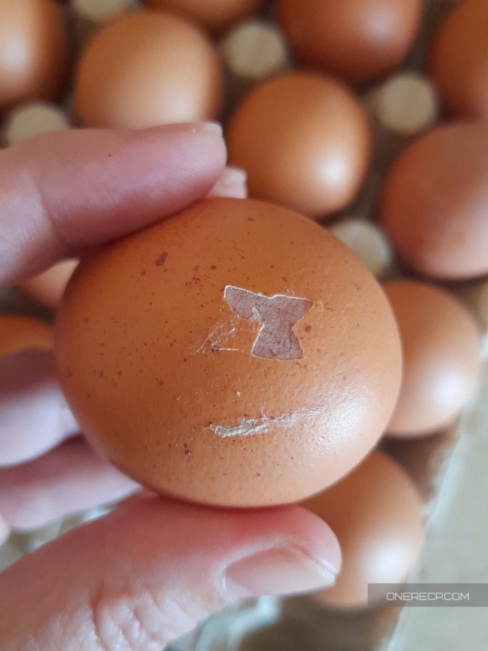 A hand holding an egg with a cracked eggshell in front of a carton with other eggs