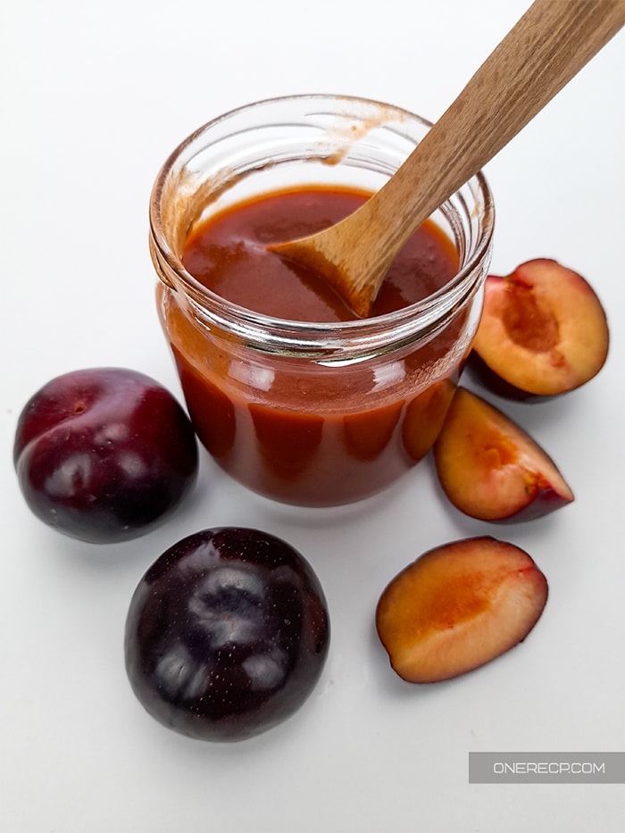 A small jar with BBQ sauce made without vinegar and a wooden spoon alongside some fresh chopped and whole plums