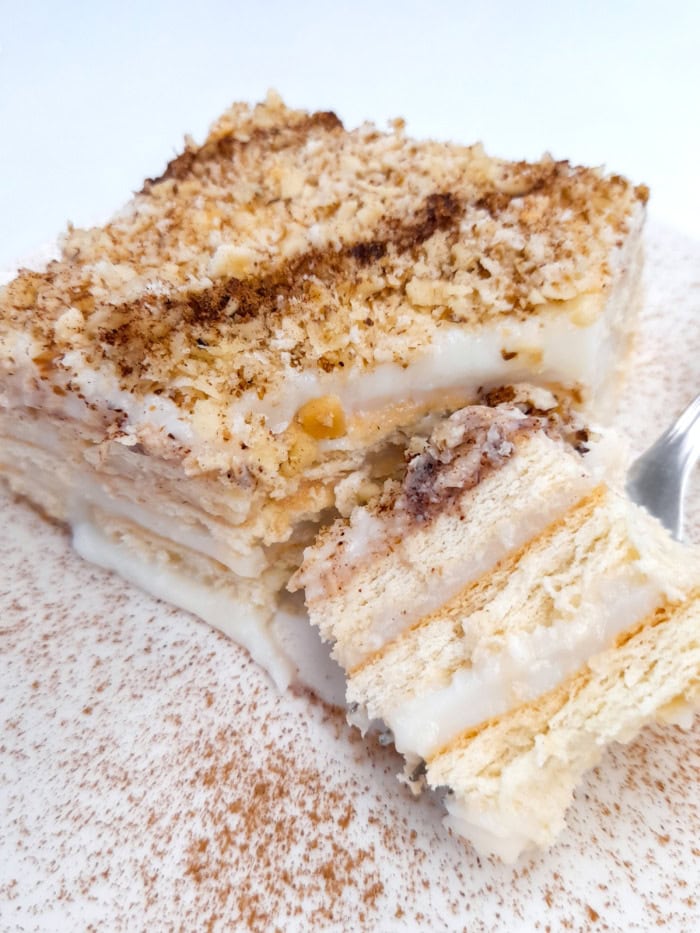 A slice of biscuit cake with layers of creamy vanilla custard, topped with crushed nuts and cinnamon