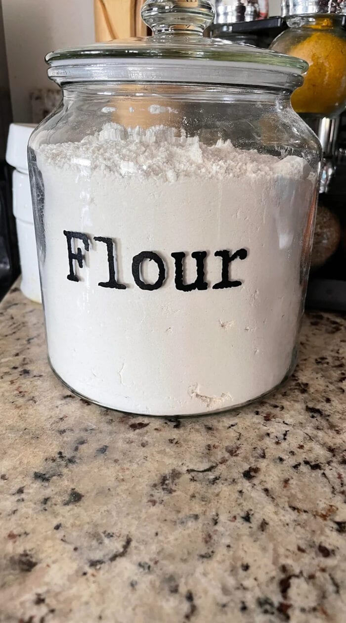 Fresh white flour stored neatly in a labeled glass jar on a kitchen counter.