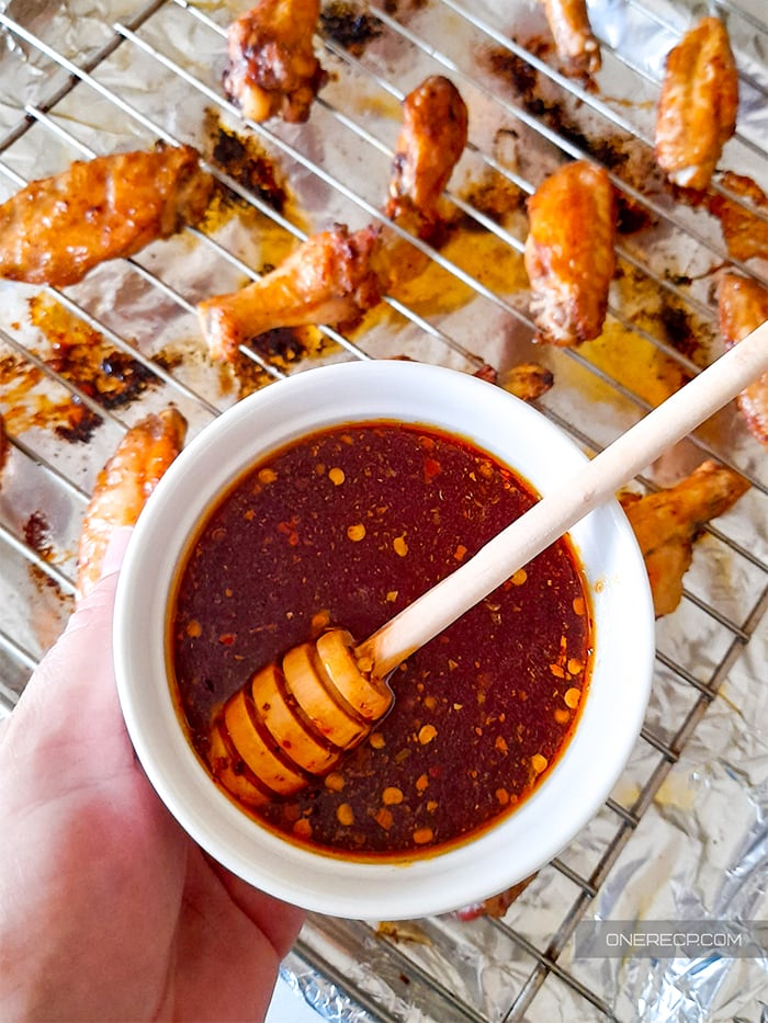 A hand holding a cup of honey hot wing sauce above some cooked chicken wings on a wire rack