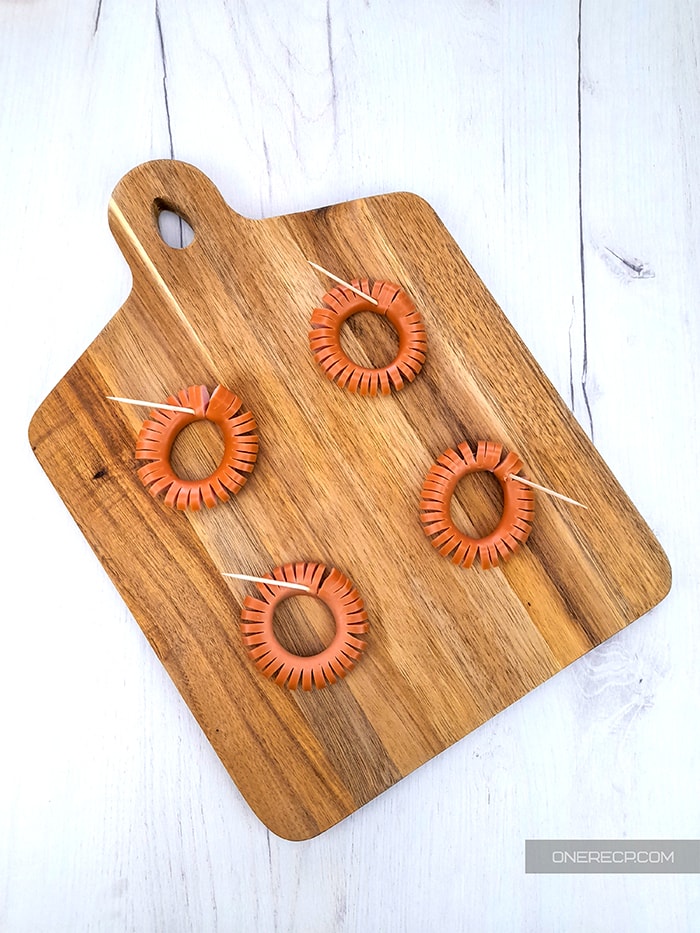 a wooden board with four wieners with equal vertical cuts held in a circular shape by toothpicks