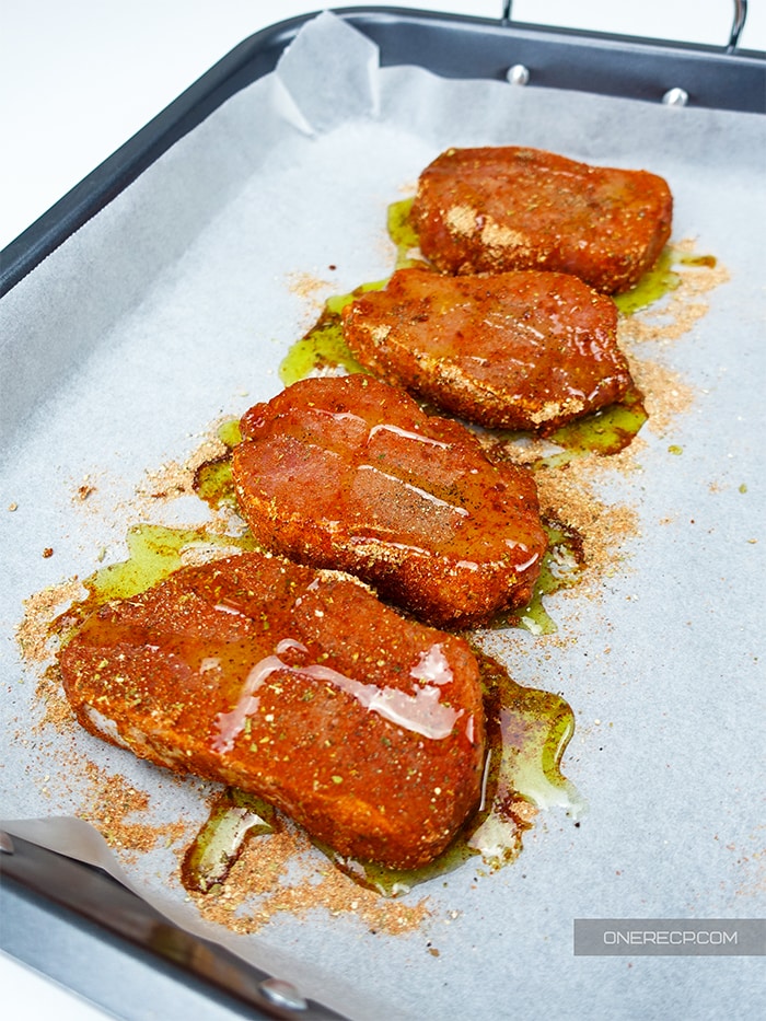 four seasoned pork chops drizzled with olive oil in a baking tray with a baking sheet