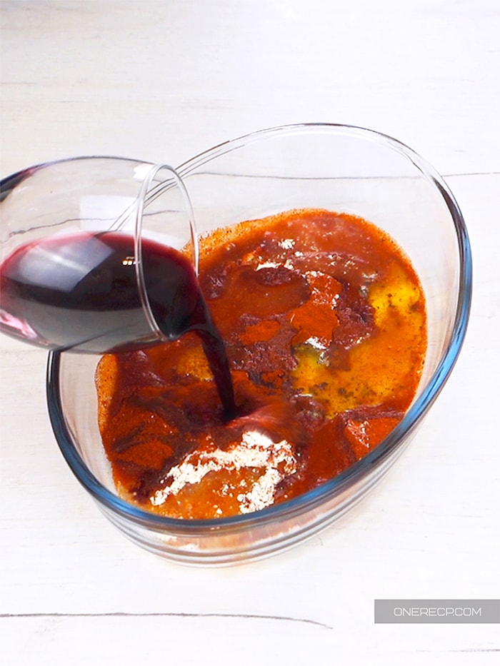 A glass of red wine being poured into a bowl with the rest of the marinade ingredients
