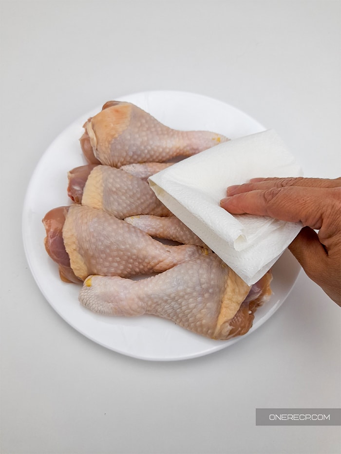 a hand removing excessive moisture from the raw chicken drumsticks by using kitchen paper