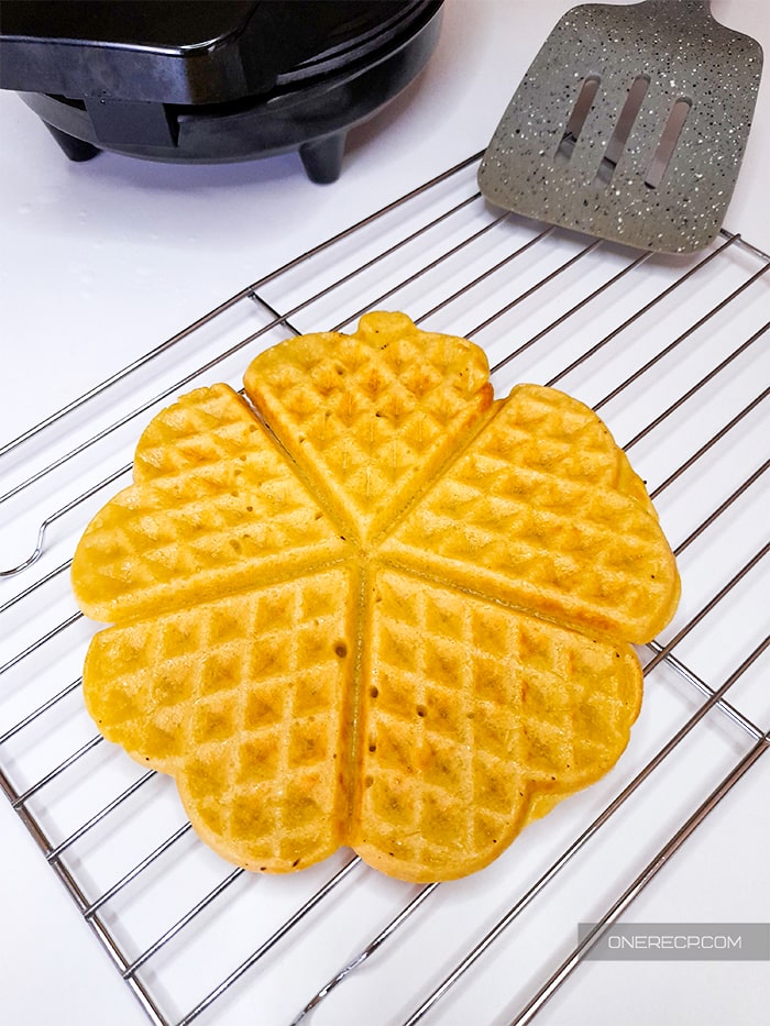 Five joined heart-shaped waffles on a wire rack with a waffle iron and a slotted turner in the distance
