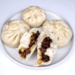 a plate of 4 pork buns with 3 being whole and one split in two so that the filling is visible
