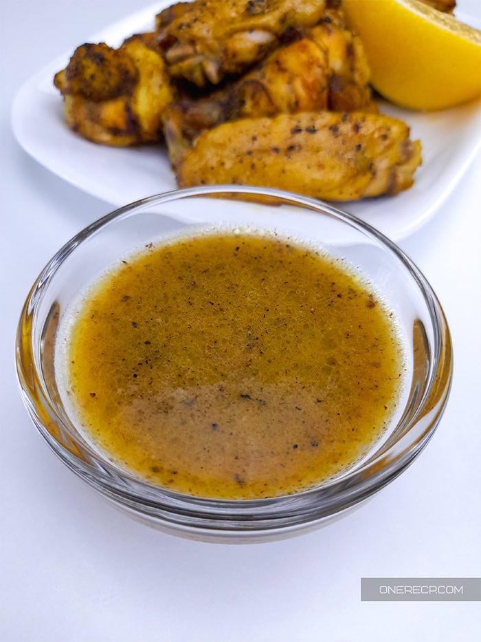 A small cup of lemon pepper sauce next to a plate with chicken wings