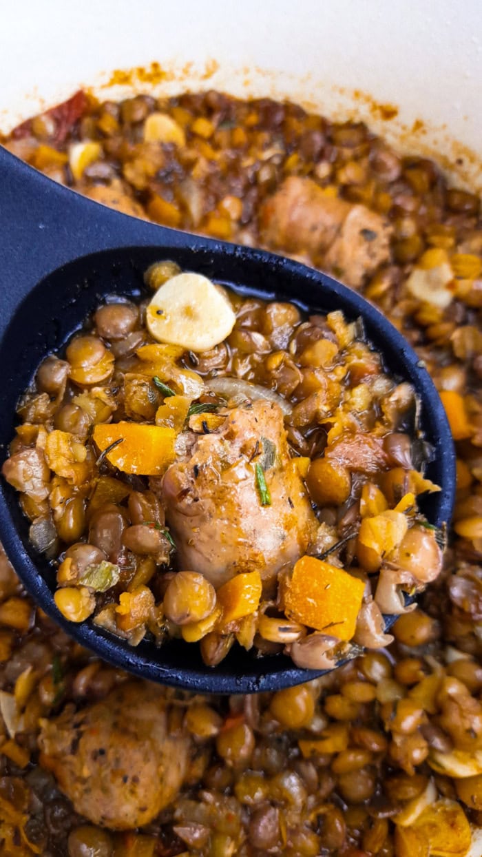 Close-up of hearty lentils and sausage stew with carrots, garlic, and herbs served in a black ladle over a pot of rustic stew.
