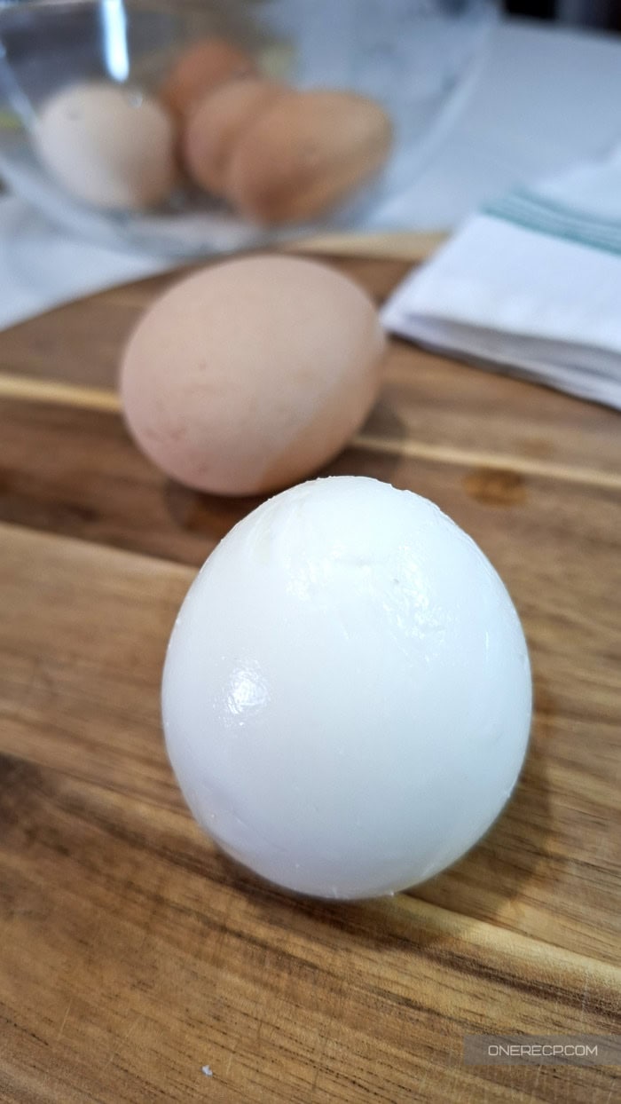 A peeled hard-boiled egg standing on a wooden cutting board with unpeeled eggs in the background.