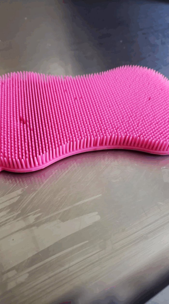 A pink silicone scrubber resting on a metal surface, demonstrating a hygienic alternative to traditional sponges.