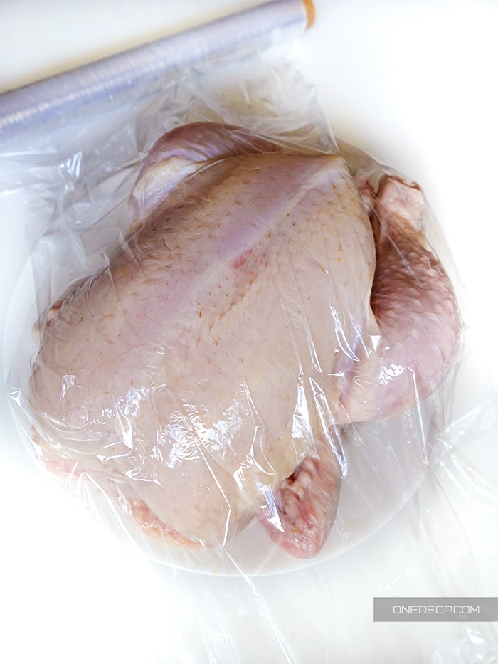 A raw chicken on a plate covered with saran plastic wrap