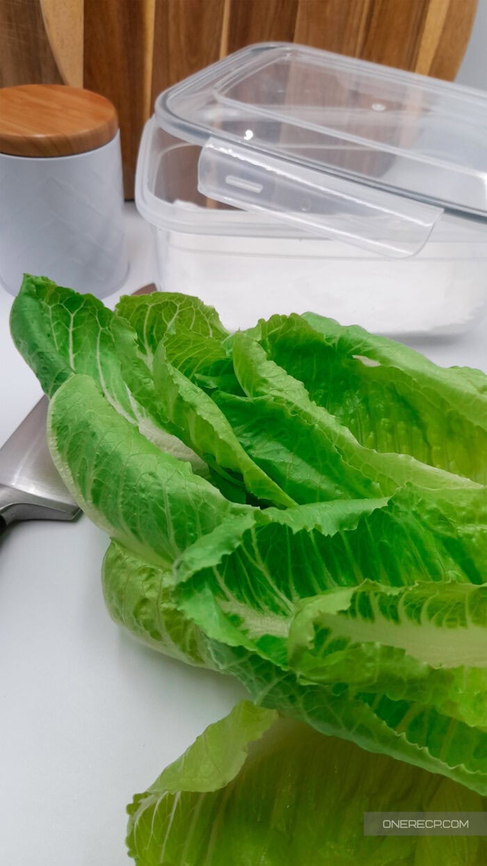 Fresh romaine lettuce leaves resting on a kitchen counter near a storage container, shown after washing and before drying to keep them crisp in the fridge.