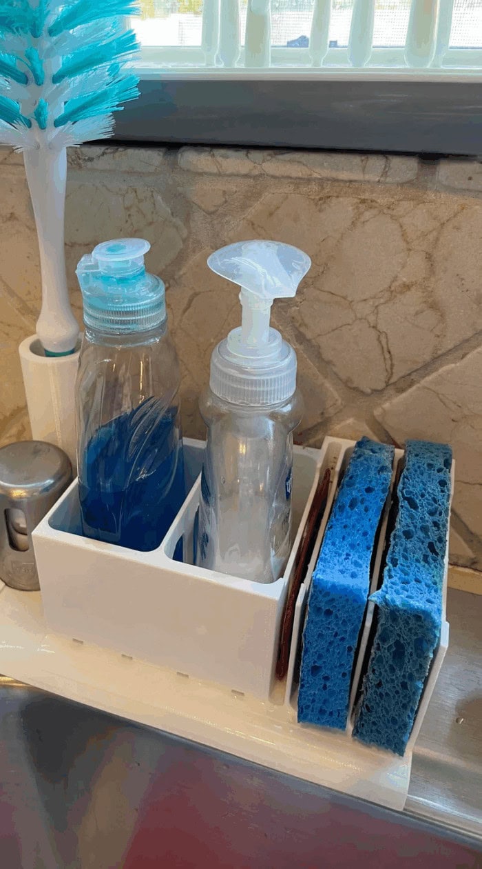Two kitchen sponges stored upright beside dish soap bottles, showing how alternating sponges helps them dry and stay odor-free.