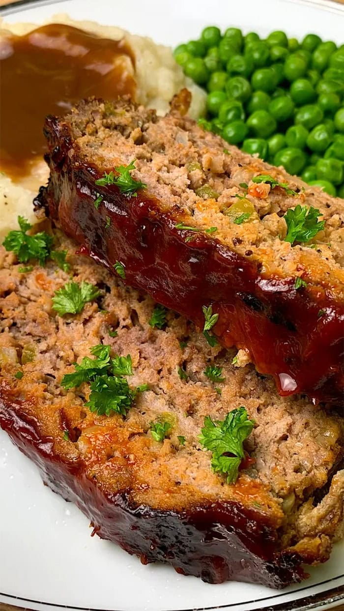 Two thick slices of glazed meatloaf served with mashed potatoes, brown gravy, and green peas on a white dinner plate.