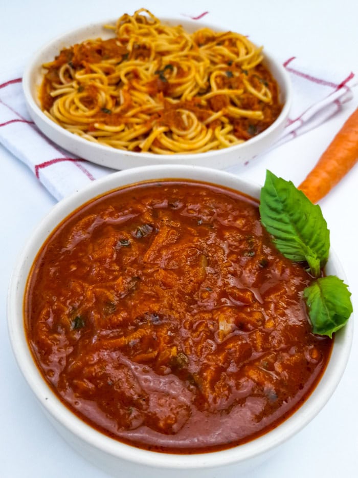 A bowl of tomato sauce with carrots garnished with basil with a plate of spaghetti in the background