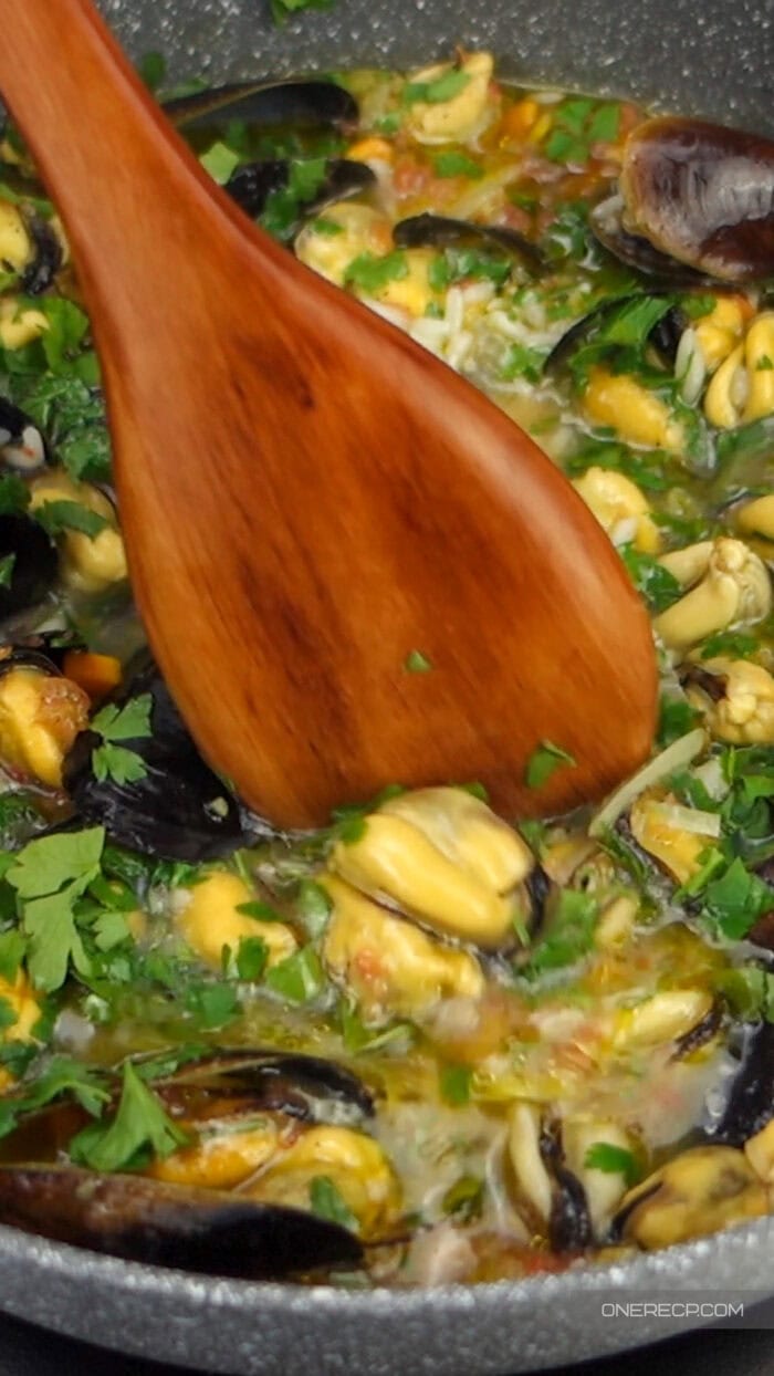 A wooden spoon stirring a seafood dish with mussels and herbs in a pan, showing its smooth seasoned surface in use.