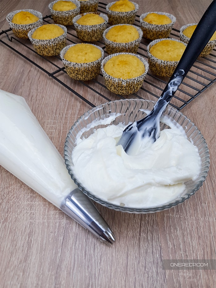 A glass bowl of vanilla frosting next to a full piping bag and a rack of cupcakes in the background