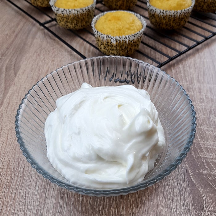 a glass bowl of fluffy vanilla frosting without butter in front of a rack with cupcakes