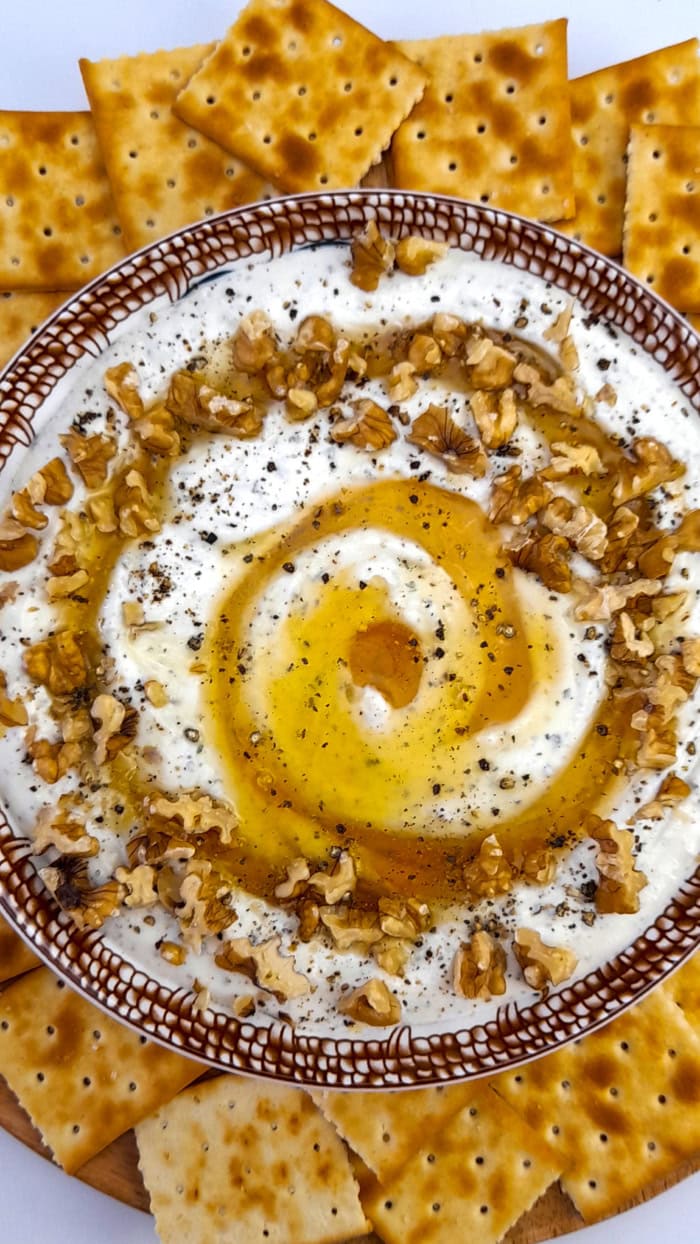 Creamy whipped feta dip drizzled with honey and olive oil, topped with walnuts and black pepper, served with crispy crackers on a wooden board.